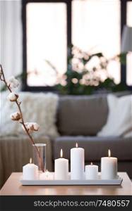 decoration, hygge and cosiness concept - burning white candles and cotton flower branch on table at cozy home. burning white candles on table at cozy home