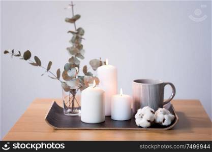 decoration, hygge and cosiness concept - burning white candles, mug, branches of eucalyptus populus and cotton flowers on table. candles, mug and branches of eucalyptus on table
