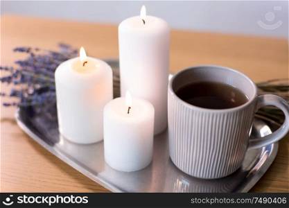 decoration, hygge and cosiness concept - burning white candles, tea in mug and lavender flowers on tray on table. candles, tea in mug and lavender flowers on table