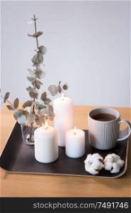 decoration, hygge and cosiness concept - burning white candles, tea in mug, branches of eucalyptus populus and cotton flowers on table. candles, tea and branches of eucalyptus on table