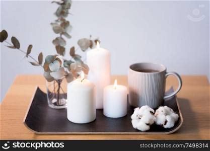 decoration, hygge and cosiness concept - burning white candles, tea in mug, branches of eucalyptus populus and cotton flowers on table. candles, tea and branches of eucalyptus on table