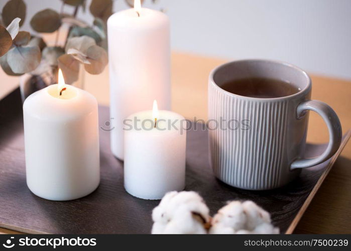 decoration, hygge and cosiness concept - burning white candles, tea in mug, branches of eucalyptus populus and cotton flowers on table. candles, tea and branches of eucalyptus on table