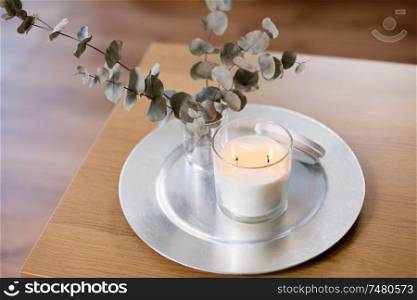 decoration, hygge and cosiness concept - burning white fragrance candle on tray and branches of eucalyptus populus on table. burning white candles on table at cozy home