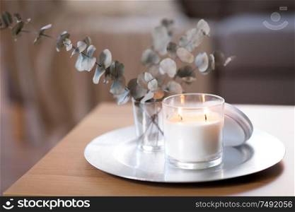 decoration, hygge and cosiness concept - burning white fragrance candle on tray and branches of eucalyptus populus on table at cozy home. burning fragrance candle on table at cozy home