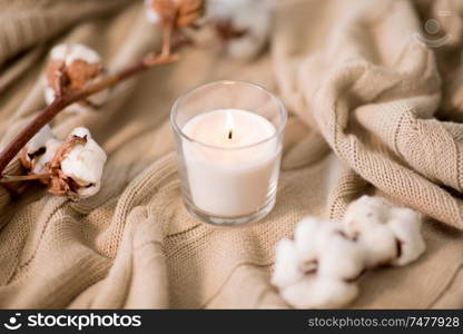 decoration, hygge and cosiness concept - burning wite fragrance candle and cotton flower on knitted blanket. burning candle and cotton flower on blanket