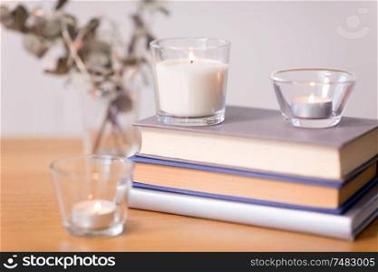 decoration, hygge and cosiness concept - fragrance candles burning in glass holders and books on wooden table. fragrance candles burning and books on table