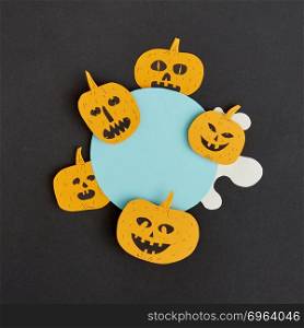 Decorative application handcraft from paper with terrible yellow pumpkins smiling on a blue round frame on a black background, place for text. Halloween concept. Flat lay.. Creative Halloween handmade blue frame with paper horrible smiling and laughing pumpkins on a black paper background.