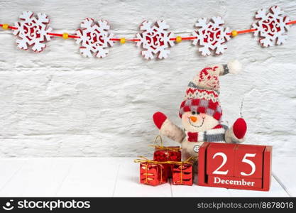 Decorative Christmas snowman, gift boxes and red wooden perpetual calendar of cube with date on white brick wall background with festive garland. Banner, header, New Year background with copy space. Decorative christmas tree, gift boxes and wooden calendar on white wooden background.
