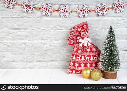 Decorative Christmas tree with two golden balls and red gift bag with christmas pattern on white brick wall background with snowflake garland. New Year background copy space, header, banner for site