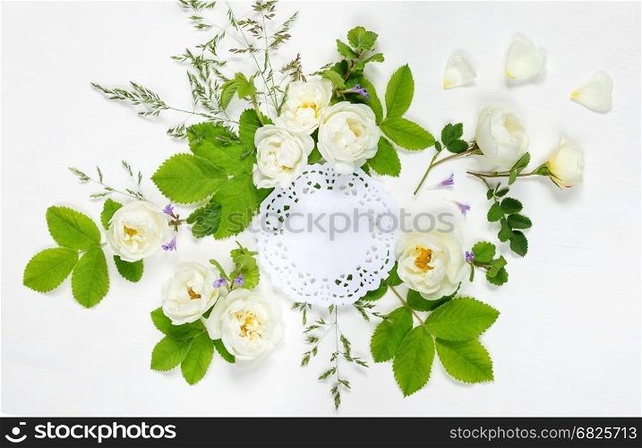 Decorative composition in retro style consisting of round white paper openwork doily and white wild rose flowers with green leaves on white background. Top view, flat lay