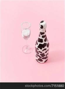 Decorative white painted champagne bottle with black spots and glass of white powder as a snow on a light millennial pink background, copy space. Congratulation card.. Holiday glass of white powder and creative painted wine bottle.