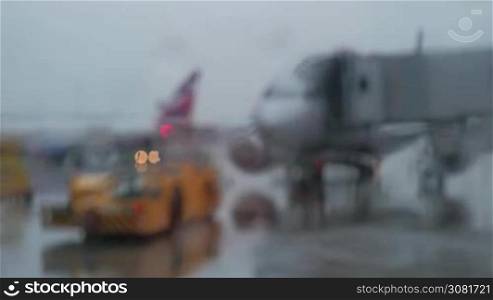 Defocused airport view through the glass on rainy day. Airplane with boarding bridge and aircraft tow tractor
