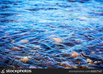 Defocused blue water surface background. Stony bottom of a clear mountain river. Flecks of sunlight and ripples on the water of river. Water background. Water surface texture.. Defocused blue water surface texture background