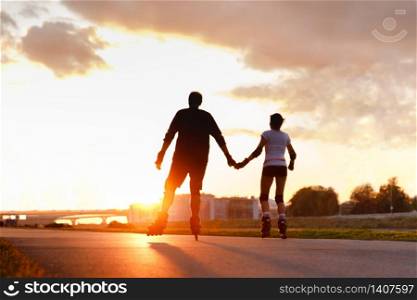 Defocused couple riding on roller skates, holding hands on the street during sunset in summer, back view from the bottom. Copy space. Active lifestyle concept.