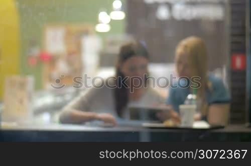Defocused shot of friendly meeting in cafe. Two women having a talk, drinking coffee and using digital tablet. View though the window in rainy evening