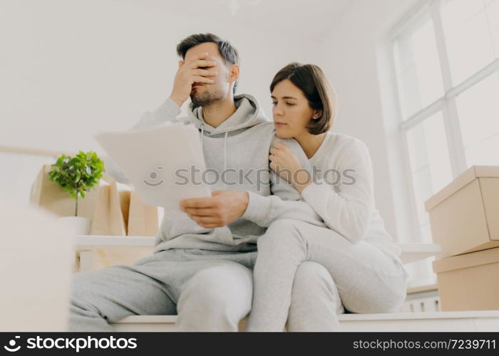 Dejected husband and wife manage finances, receive bill, face financial problem, have gloomy expressions, sit together in empty room, big window behind, cardboard boxes with personal stuff near