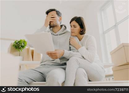 Dejected husband and wife manage finances, receive bill, face financial problem, have gloomy expressions, sit together in empty room, big window behind, cardboard boxes with personal stuff near