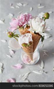 Delicate pink and white peony flowers in a wafer cones in a glass standing on a gray stone table with petals on it, copy space. Concept of congratulations for Valentines Day.. Greeting card with three sweet wafer cones and gentle peony flowers in a glass vase, petals on a gray concrete background.