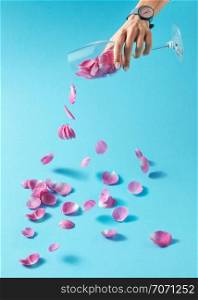 Delicate pink rose petals are pouring from transparent glass which girl&rsquo;s hand holds with wirstwatch on it. Concept production of wine from natural products.. Woman&rsquo;s hand with watch is pouring petals of pink rose from glass on a blue background. Place for text.