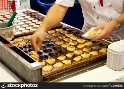 Delicious authentic Dutch food from The Netherlands called poffertjes, which are mini pancakes, being prepared on a typical hot plate while cook is collecting them to sell.