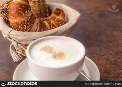 Delicious breakfast with a cup of milk coffee and fresh, sesame croissants in a rustic wicker basket, on a brown table.