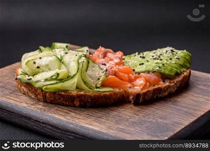 Delicious brown bread toast with salmon, avocado, cucumber and sesame seeds on a textured concrete background
