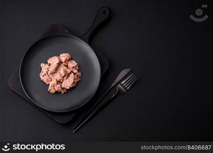 Delicious can≠d tuna meat on a black ceramic plate on a dark concrete background. Hea<hy food pre¶tion