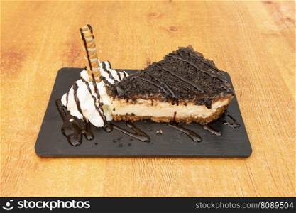 Delicious cheesecake with pieces of oreo, chocolate syrup, cream and a waffle on a black slate plate