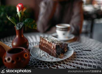 Delicious chocolate cake and a cup of coffee on table in cafe. Table decorated with white knitted tablecloth and vase with rose. Woman sits at table in cafe, selective focus.. Delicious chocolate cake and a cup of coffee