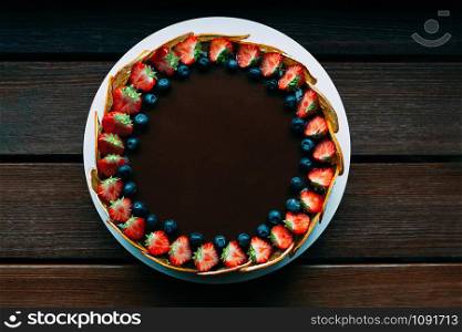 Delicious chocolate cake decorated with fresh strawberries, blueberries, mint, and candied oranges on the brown wooden table. Luxurious dark glaze. Image for menu or confectionery catalog