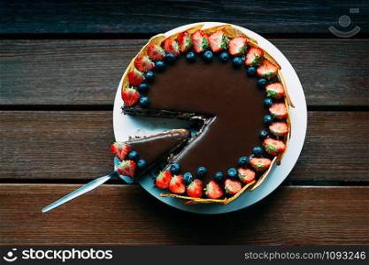 Delicious chocolate cake decorated with fresh strawberries, blueberries, mint, and candied oranges on wooden table background. Luxurious dark glaze. Image for menu or confectionery catalog