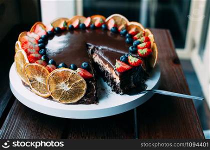 Delicious chocolate cake decorated with fresh strawberries, blueberries, mint, and candied oranges on the brown wooden table. Luxurious dark glaze. Image for cafe menu or confectionery catalog