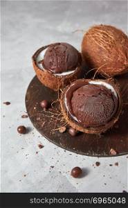 Delicious chocolate sorbet in a coconut shell on a wooden board on a gray concrete background with place for text. Vegetarian concept of diet eating. Fresh appetizing chocolate ice cream in a coconut shell with chocolate balls on a wooden board on a gray.