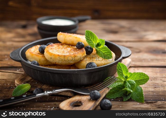 Delicious cottage cheese pancakes or syrniki with fresh blueberry in cast-iron pan on dark wooden rustic background, above view. Tasty breakfast.