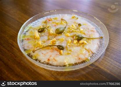 Delicious dish with prawn carpaccio garnished with capers and nut sauce served on plate on wooden restaurant kitchen. Exquisite dish with prawn carpaccio garnished with capers and nut sauce