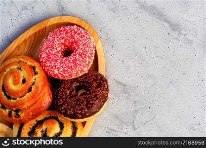 Delicious donuts with chocolate and strawberry icing and buns with poppy seeds and freshly baked raisins, close-up on an oval wooden tray and a gray concrete surface. Pastry sweets for breakfast, image with copy space.. Delicious chocolate donuts and buns with poppy seeds and raisins lie on an oval wooden tray and a gray concrete surface.