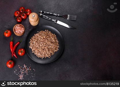 Delicious fresh boiled buckwheat porridge with vegetables and spices on a black plate against a dark concrete background. Delicious fresh boiled buckwheat porridge with vegetables and spices on a black plate