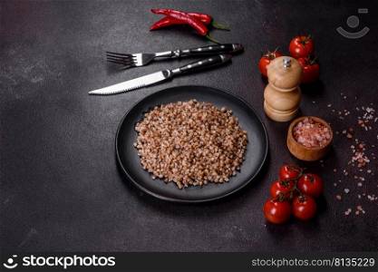 Delicious fresh boiled buckwheat porridge with vegetables and spices on a black plate against a dark concrete background. Delicious fresh boiled buckwheat porridge with vegetables and spices on a black plate