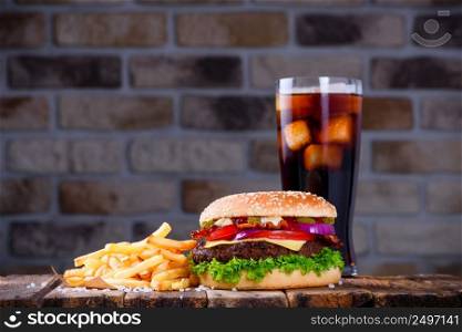 Delicious fresh hamburger with french fries and cola on wooden table. Big burger in classic american style with hot grilled patty with melted cheese on top, tomato, onion, sauces and fried chips served with cold soft drink.