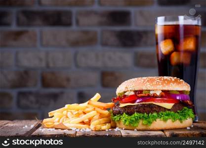 Delicious fresh hamburger with french fries and cola on wooden table. Big burger in classic american style with hot grilled patty with melted cheese on top, tomato, onion, sauces and fried chips served with cold soft drink.