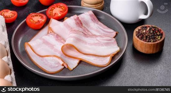 Delicious fresh raw bacon cut with slices on a grey plate against a dark concrete background. Making a delicious nutritious breakfast. Delicious fresh raw bacon cut with slices on a grey plate against a dark concrete background