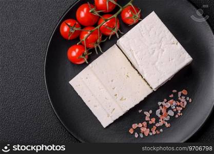 Delicious fresh ricotta cheese cut into slices with vegetables, salt and spices on a dark concrete background