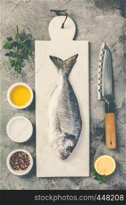 Delicious fresh sea bream fish on marble board, herbs and spices over grey stone background. Cooking concept. Delicious fresh sea bream fish, flat lay, top view