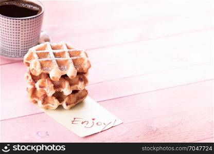 Delicious fresh waffles covered in powdered sugar, a cup of coffee and a piece of paper with the message enjoy, on a pink wooden table