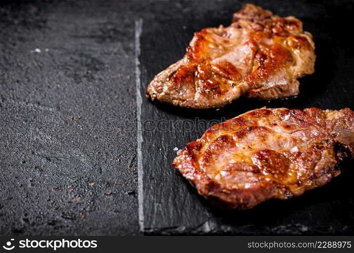 Delicious grilled pork steak on a stone board. On a black background. High quality photo. Delicious grilled pork steak on a stone board.