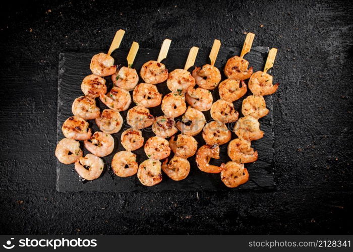 Delicious grilled shrimp on skewers on a stone board. On a black background. High quality photo. Delicious grilled shrimp on skewers on a stone board.