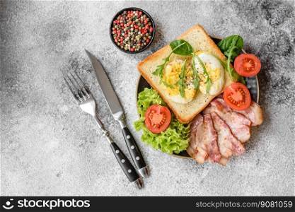 Delicious healthy lunch consisting of bacon, toast, eggs, tomatoes, lettuce and greens on a black plate on a concrete background