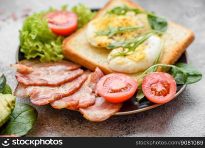 Delicious healthy lunch consisting of bacon, toast, eggs, tomatoes, lettuce and greens on a black plate on a concrete background