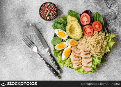 Delicious healthy lunch consisting of chicken, avocado, quinoa, eggs, tomatoes, lettuce and greens on a black plate on a concrete background