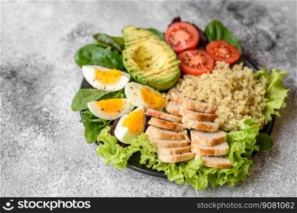 Delicious healthy lunch consisting of chicken, avocado, quinoa, eggs, tomatoes, lettuce and greens on a black plate on a concrete background